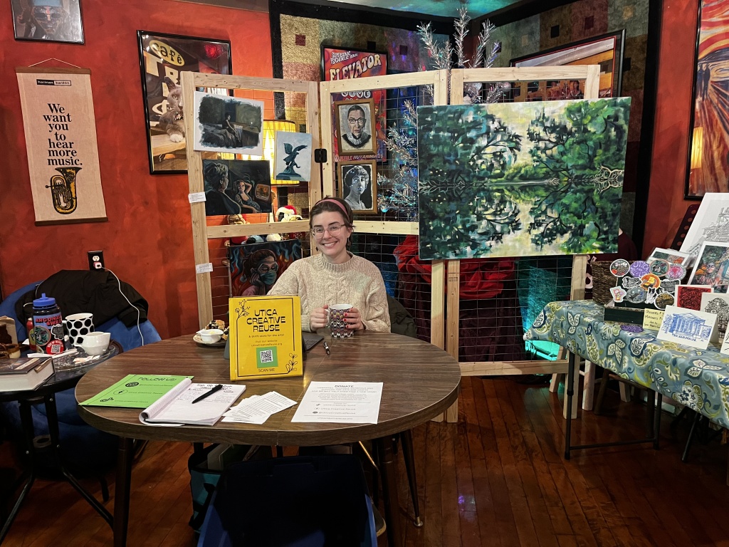Photo of Sarah Marris-Swann sitting at a table with a Utica Creative Reuse sign and informational materials laid out. In the background, pieces of artwork for sale are displayed. 